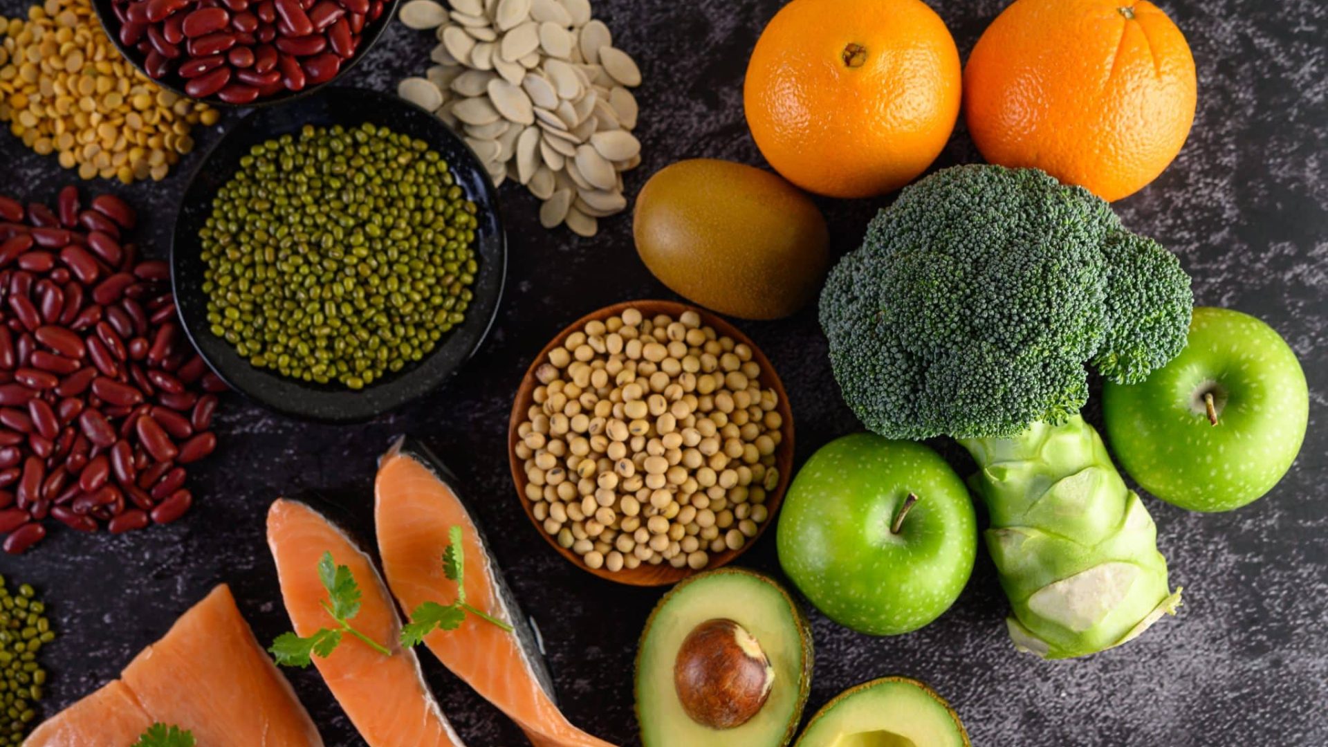 Legumes, broccoli, fruit, and salmon placed on a black cement floor. Top view.
